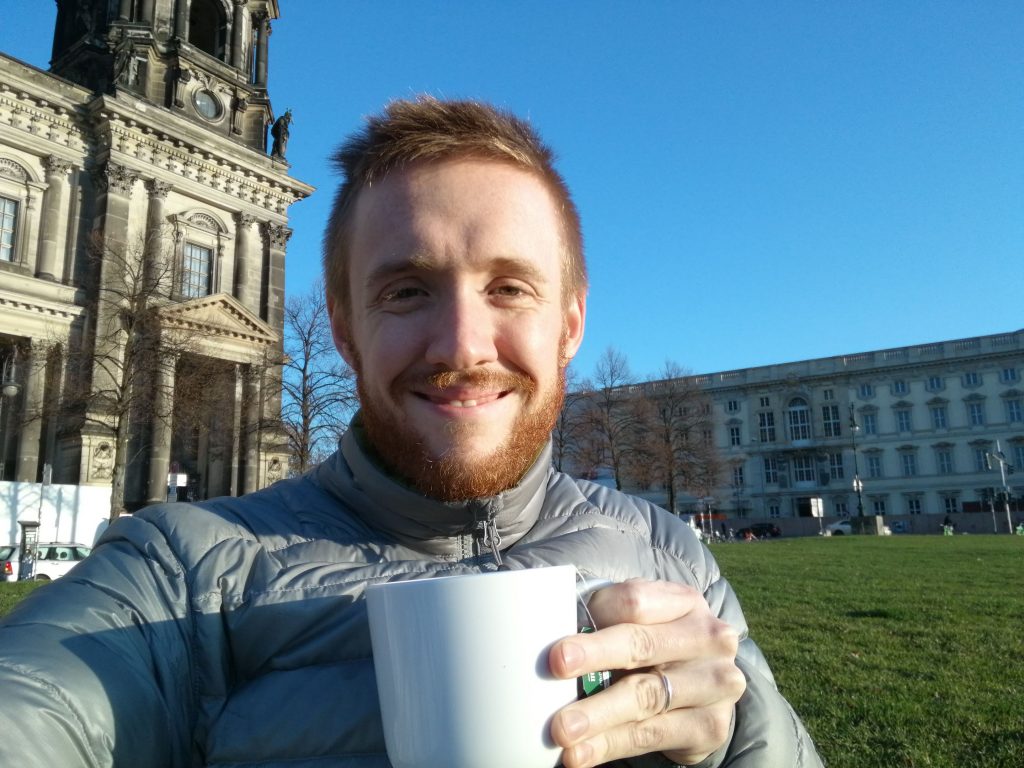 a man in a down coat smiling at the camera, drinking a cup of tea in a park. an ornamental church is in the background.