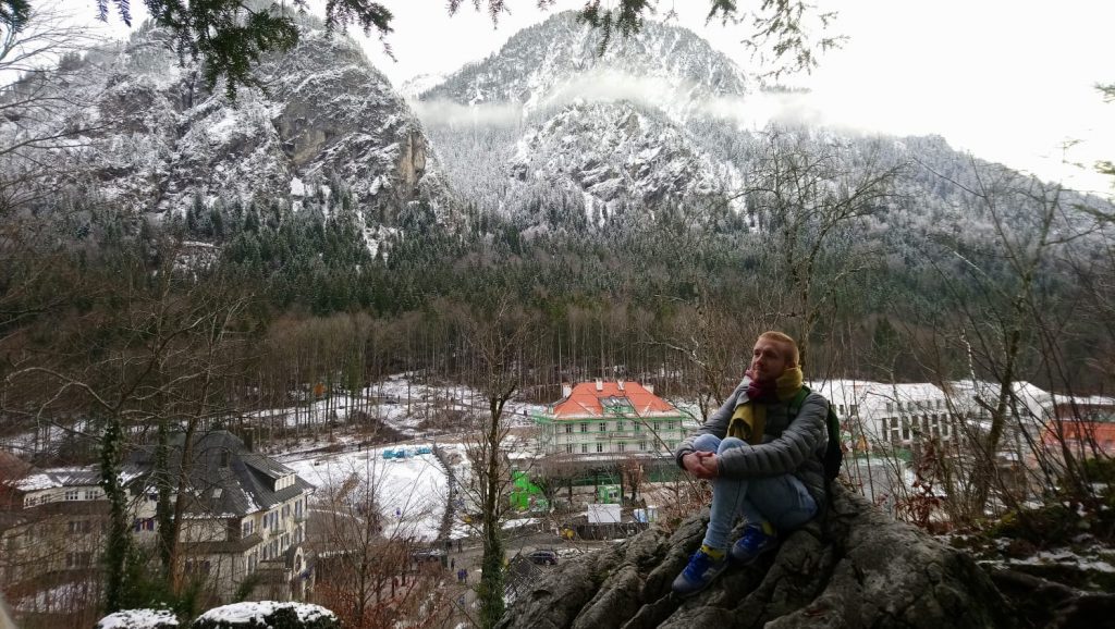 me sitting on a rock in front of a valley. there are a few scattered buildings in the valley, and snow-covered, rocky mountains rise up beyond.